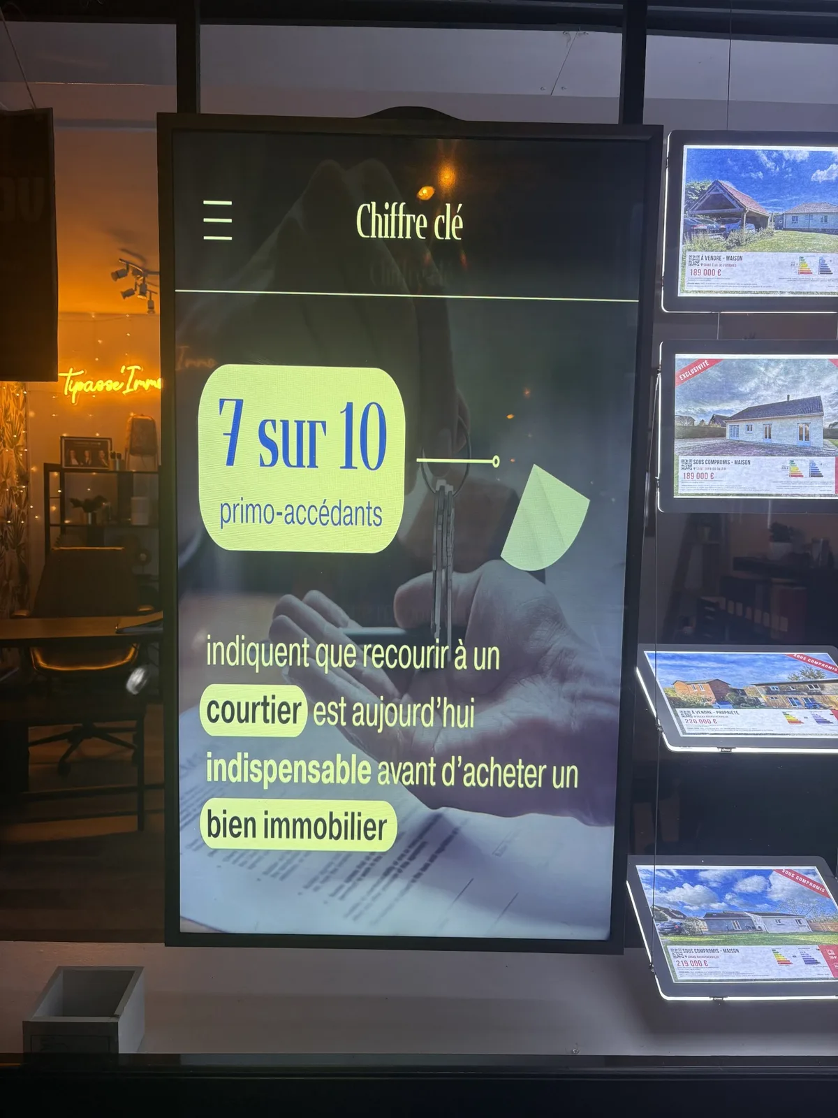 Écran en vitrine d'une agence immobilière diffusant une infographie sur le recours à un courtier