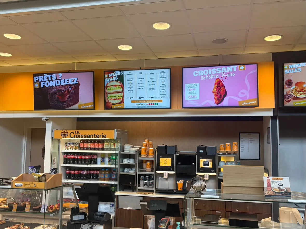 Mur de quatre écrans menu board au-dessus du comptoir La Croissanterie, affichant les menus salés, un fondant au chocolat et le croissant Octobre Rose