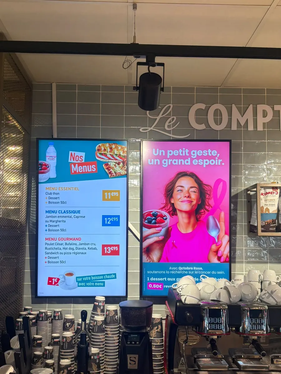 Deux écrans verticaux fixés au mur derrière le comptoir d'un café, affichant les formules sandwich, dessert et boisson et une campagne Octobre Rose